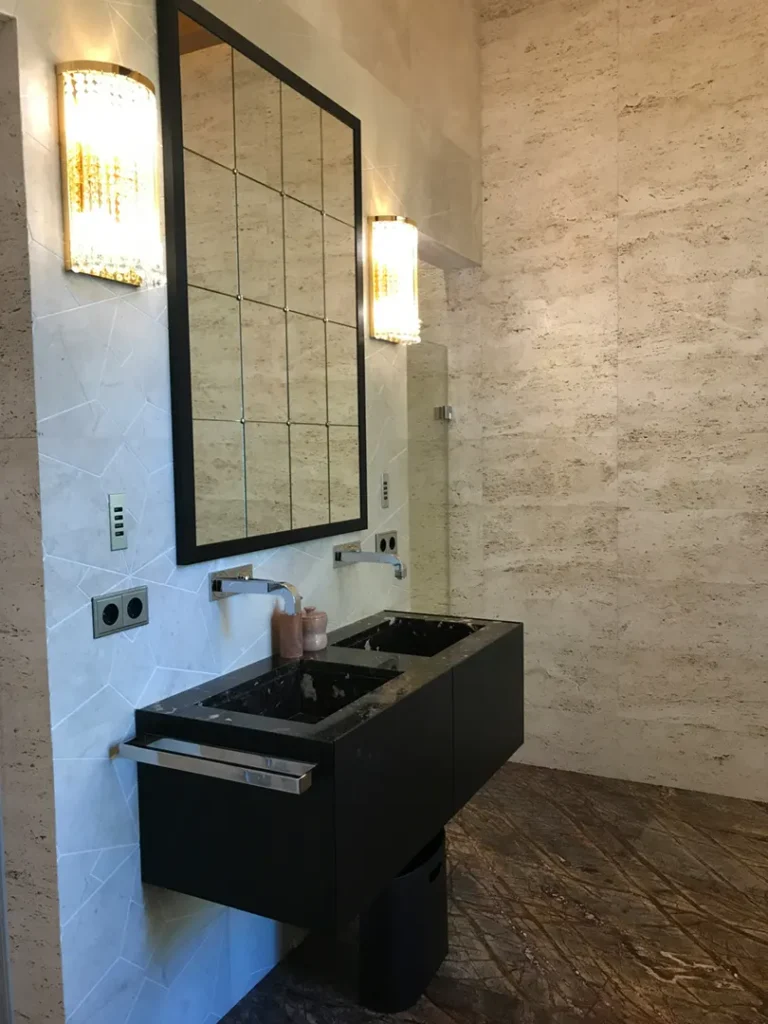 Un moderno mobile bagno su misura realizzato in legno chiaro con lavabo, specchio a parete illuminato e mobiletto sottolavabo con cassetti e ante. Il mobile bagno è completato da faretti a LED e da una finitura elegante e raffinata. La foto mostra un bagno luminoso e spazioso, con un pavimento in gres porcellanato effetto marmo e pareti in marmo grigio.